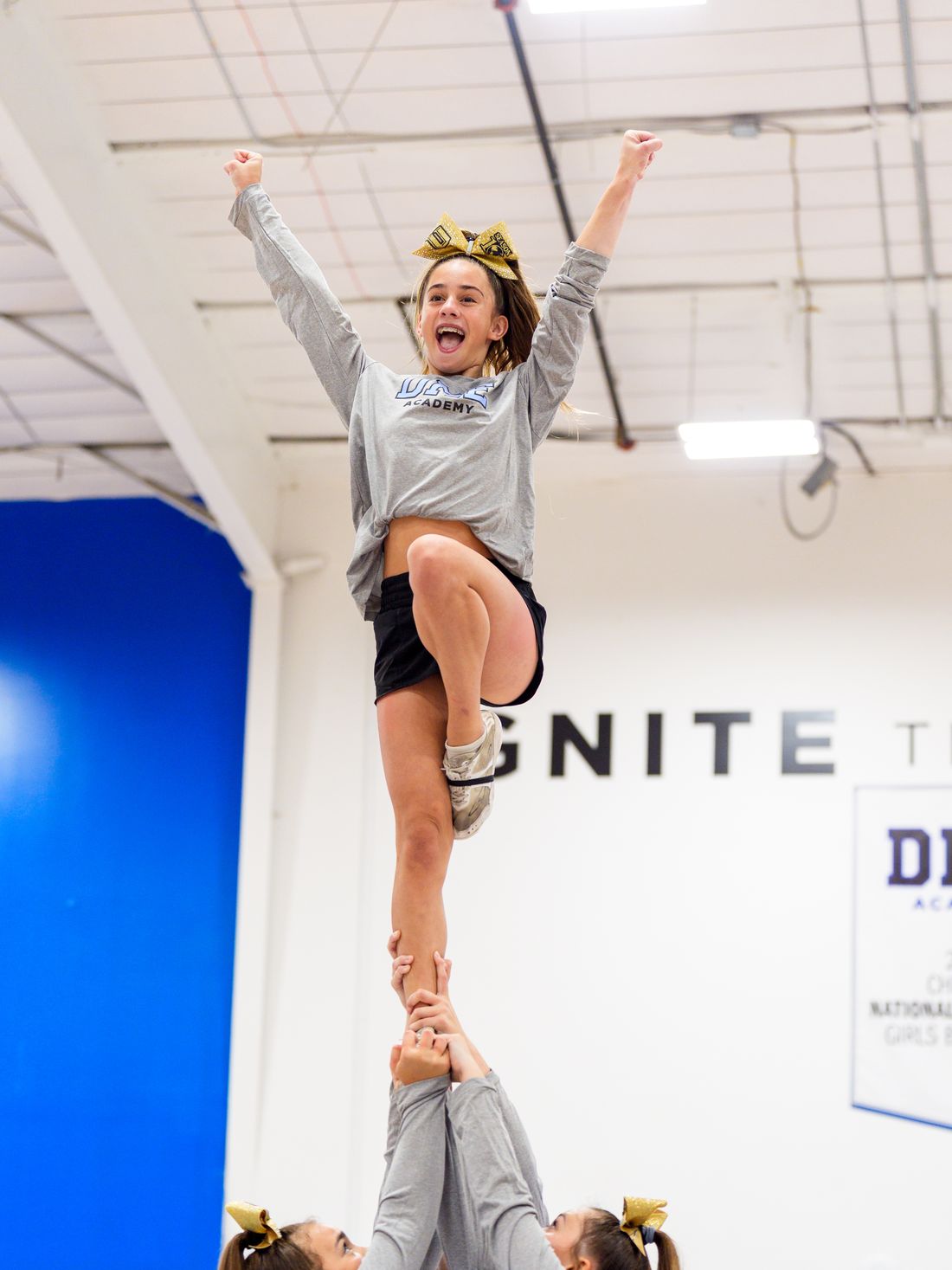 A DME cheer flyer at full extension, arms raised, beneath the Ignite The Next wall.